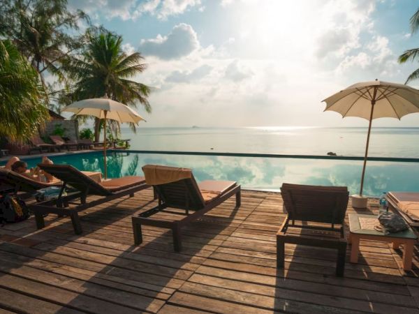 Poolside deck with lounge chairs and umbrellas facing a serene ocean, palm trees, and a tranquil sky.