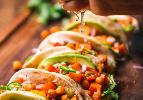 A hand squeezes lime over a row of tacos filled with chickpeas, avocado, and vegetables on a wooden board.