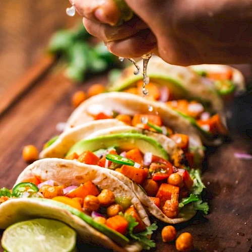 A hand squeezes lime over a row of tacos filled with chickpeas, avocado, and vegetables on a wooden board.