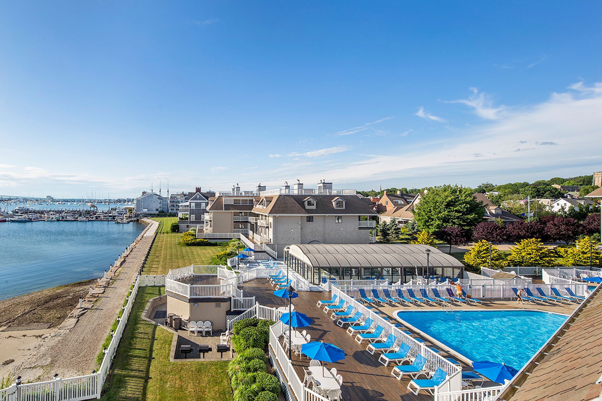 A coastal resort with a large pool, blue umbrellas, and sun loungers, next to a calm waterfront and a clear blue sky.