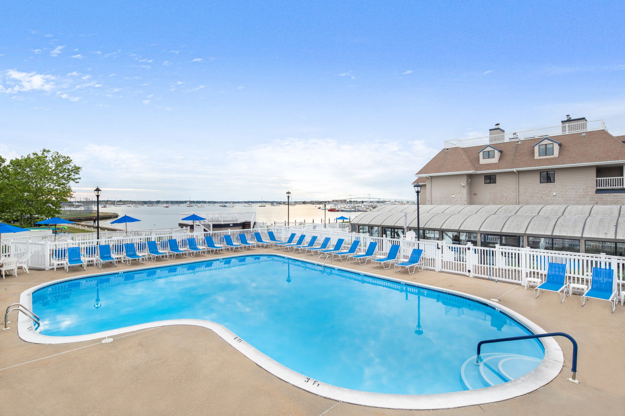 A pool with blue lounge chairs by a building, overlooking a scenic waterfront view under a clear blue sky.