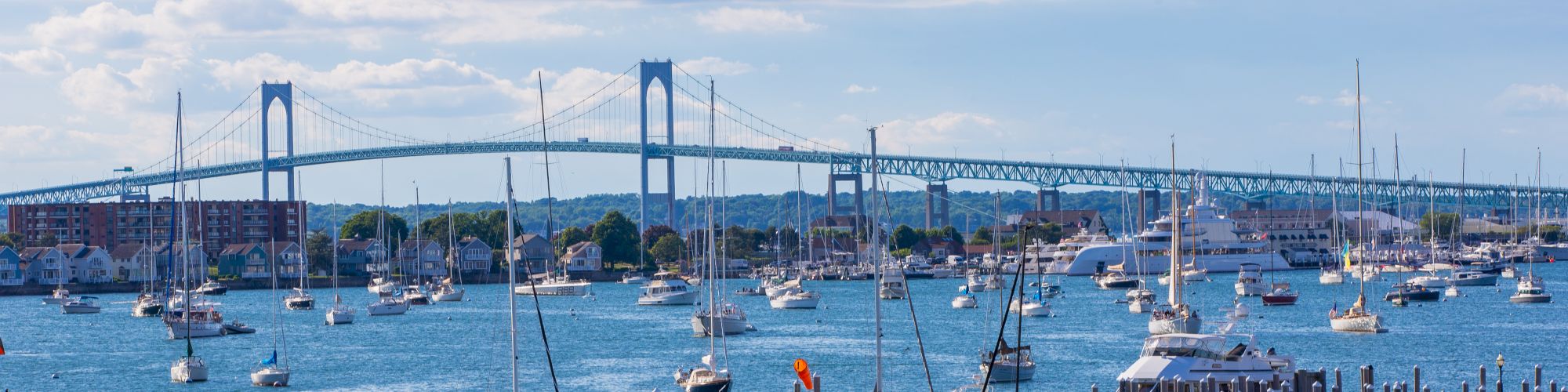 A scenic view of a marina with numerous boats, calm blue water, and a large bridge in the background under a partly cloudy sky.