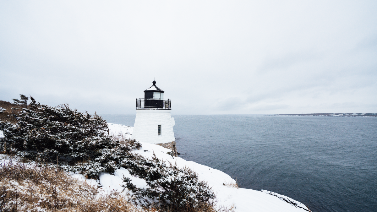 A snowy coastal scene with a white lighthouse overlooking the ocean, surrounded by shrubs and grass.