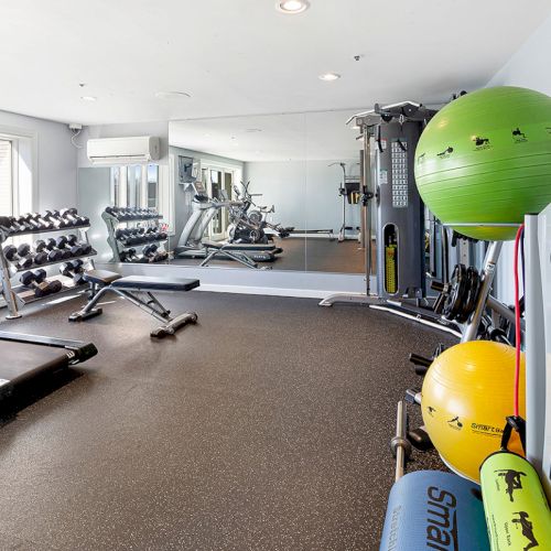 A modern gym with exercise equipment, including treadmills, weights, yoga balls, and resistance trainers. Mirrors line one wall.