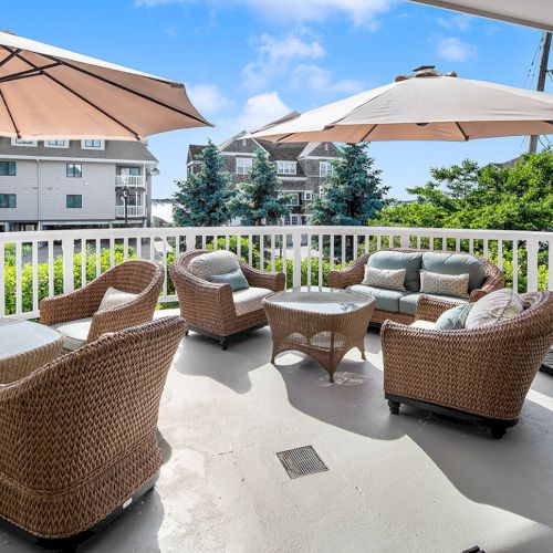 Outdoor patio with wicker furniture, tables, and umbrellas. Surrounded by white railing, greenery, and buildings in the background.