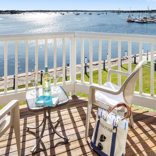 A deck with chairs, a table holding a bottle and glass, overlooking a calm body of water and boats in the distance on a sunny day.