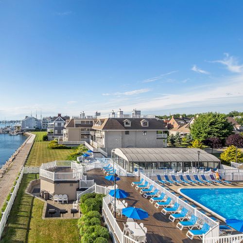 A coastal resort with a swimming pool, sun loungers, and blue umbrellas by the water, surrounded by greenery and buildings.