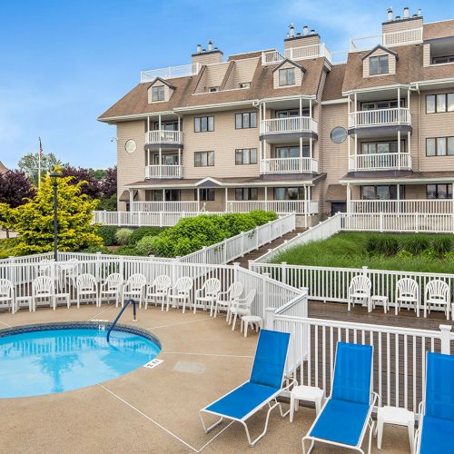 The image shows a hotel with balconies, a small pool, and surrounding blue lounge chairs, enclosed by a white fence under a clear sky.