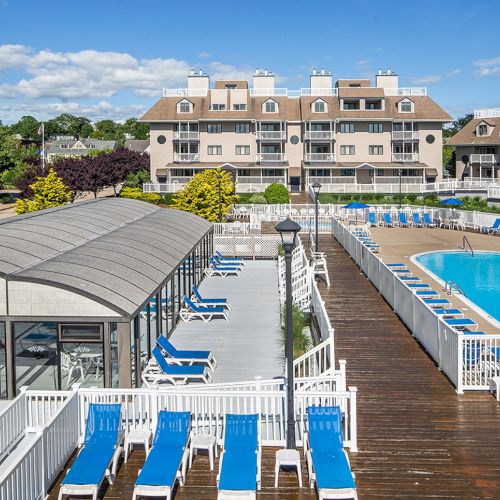The image shows a resort with a swimming pool, lounge chairs, a covered area, and surrounding buildings under a blue sky.