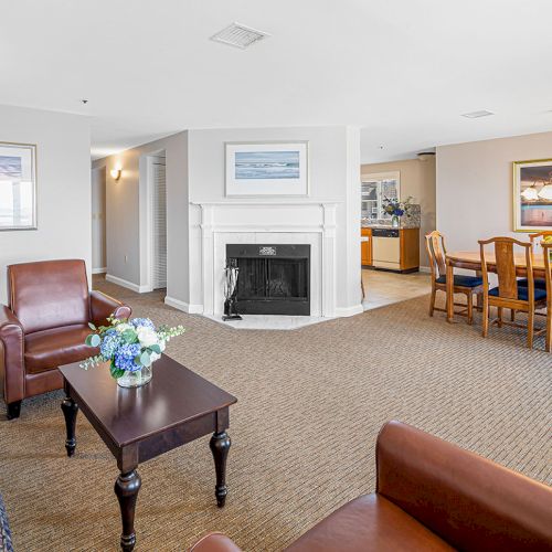 The image shows a spacious living room with a fireplace, leather chairs, a dining table, and paintings on the walls, providing a cozy atmosphere.