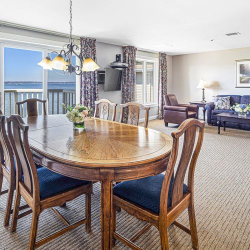 A spacious living and dining area with a wooden table, chairs, sofas, and large windows showing an ocean view. Bright and inviting space.
