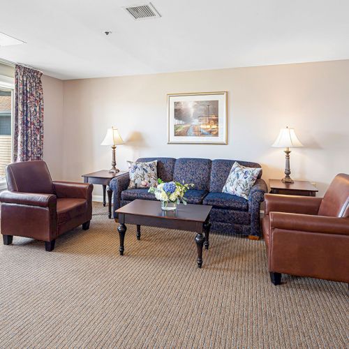 A cozy living room with a couch, two chairs, a coffee table, and table lamps, overlooking a body of water through a large window.
