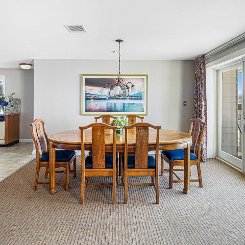 The image shows a dining room with a wooden table and chairs, a painting on the wall, and glass doors leading to a balcony.