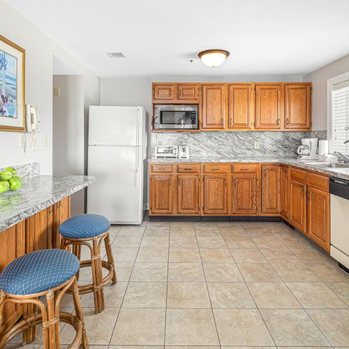 A kitchen with wooden cabinets, a fridge, dishwasher, countertop with green apples, stools, and a vase with flowers on the right.