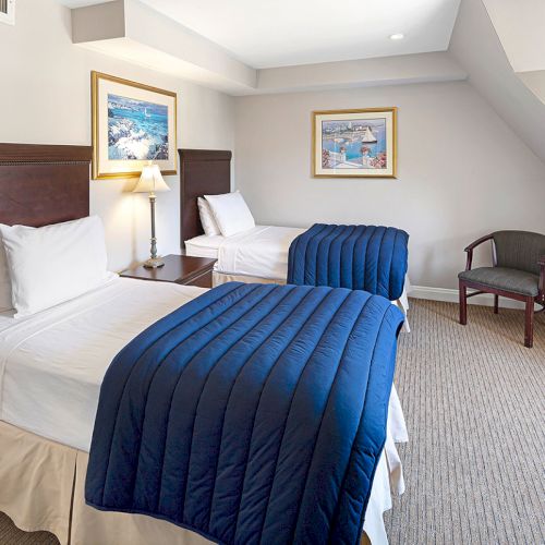 The image shows a bedroom with two single beds, blue comforters, a lamp, two framed pictures on the wall, and a chair in the corner.