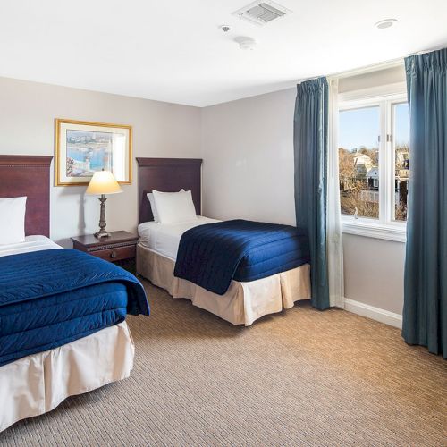 A cozy room with two single beds, blue bedding, a lamp, a window with curtains, a framed picture, and an armchair in the corner.