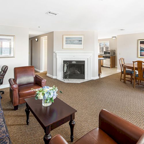 The image shows a living room with a fireplace, sofa, armchairs, coffee table, and adjoining dining area, decorated with framed artwork and flowers.