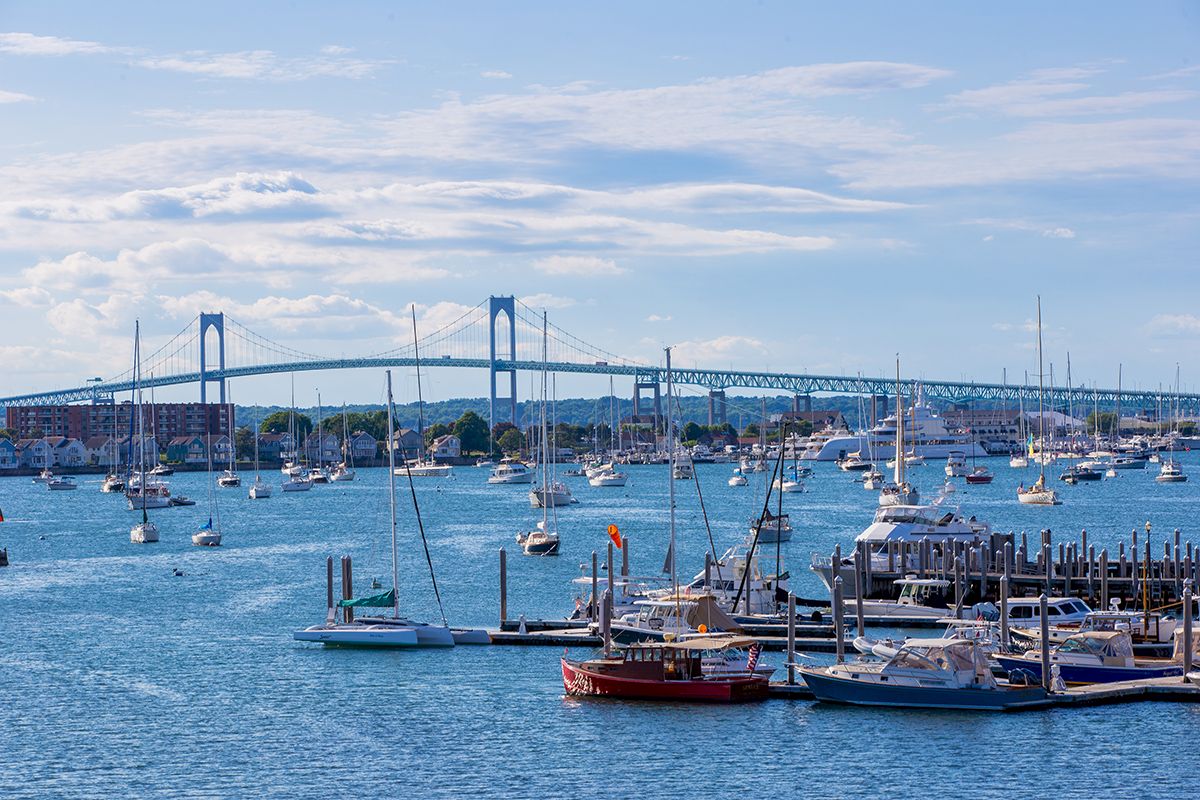 The image shows a harbor with docked boats and sailboats on water, a large bridge in the background, under a partly cloudy sky.