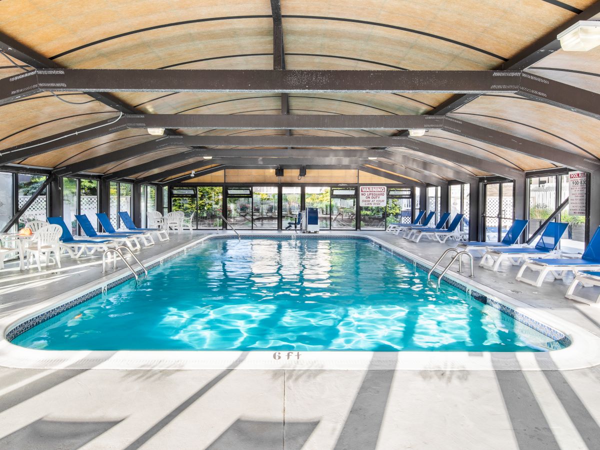 The image shows an indoor swimming pool with blue lounge chairs on each side and a transparent, curved roof.