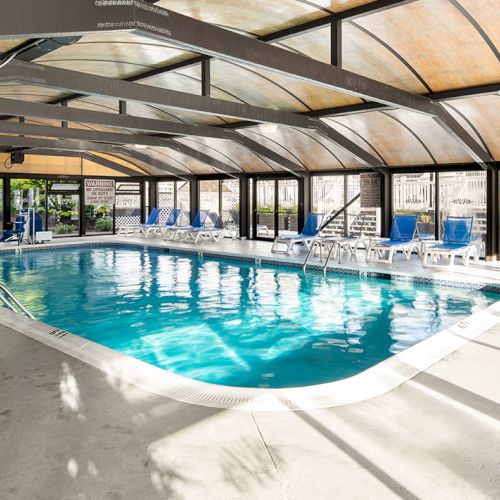 An indoor swimming pool with clear water, surrounded by blue lounge chairs and enclosed by a glass and metal structure for natural lighting.