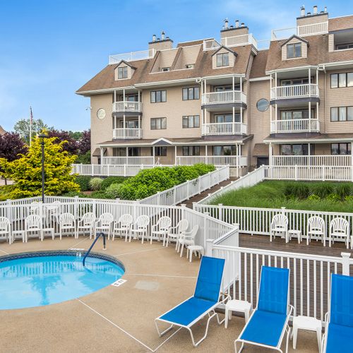 The image shows a multi-story building with balconies, a fenced circular pool, and blue lounge chairs on a patio. The sky is clear.
