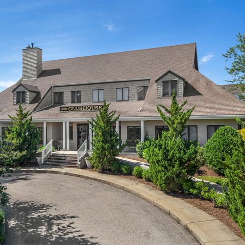 The image shows a large, two-story building with a sloped roof, surrounded by trees, with a driveway leading to the entrance.