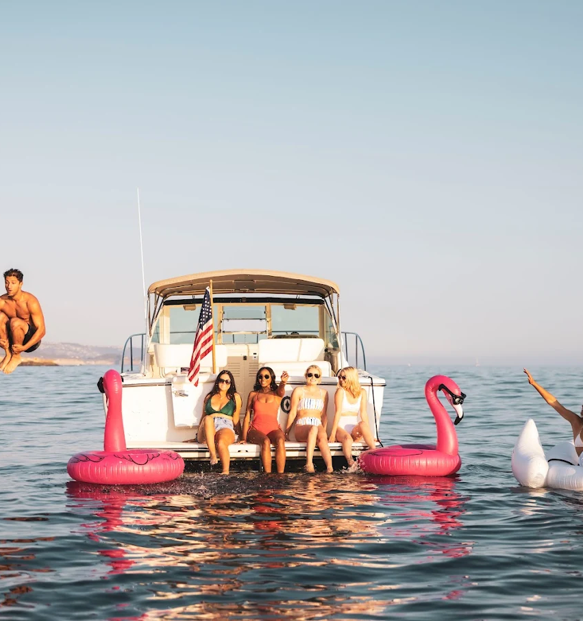 A group of friends on a boat with pink flamingo floaties, two people on a wingboard/skimming near the water, and a bright sunny day.
