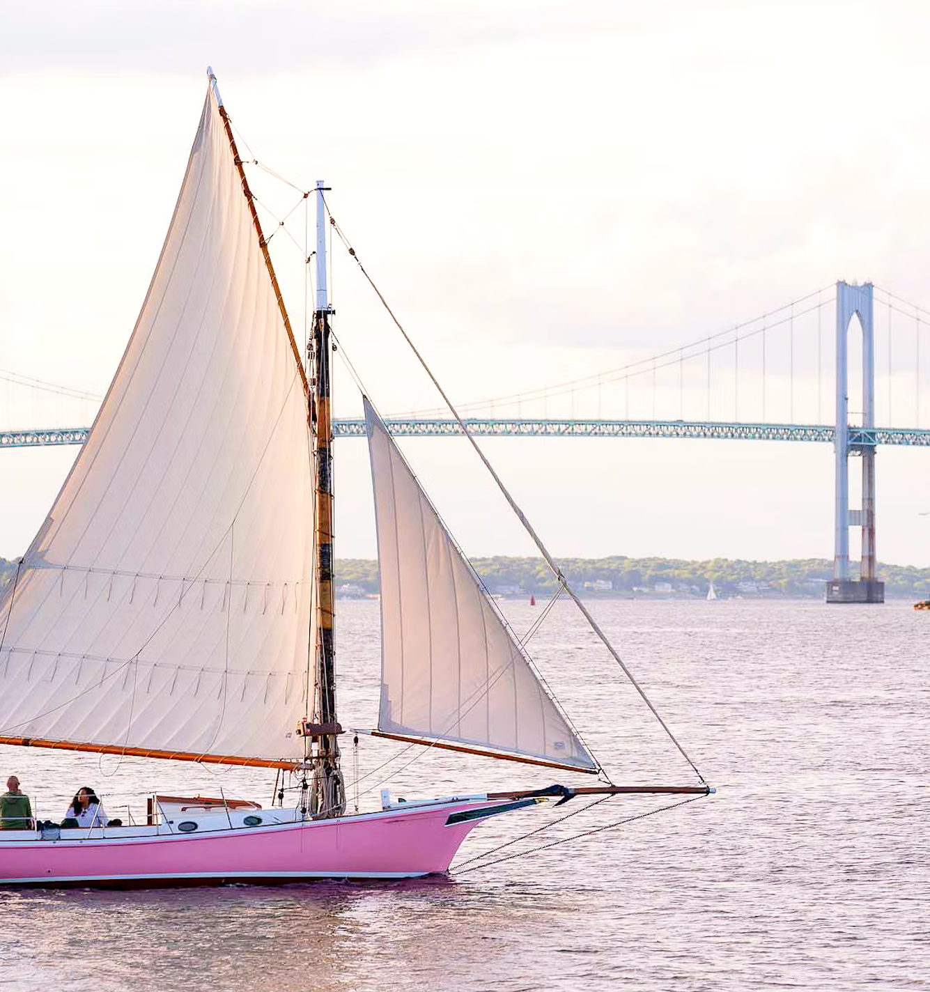 A pink sailboat with white sails cruises on a river near a large suspension bridge under a cloudy sky.