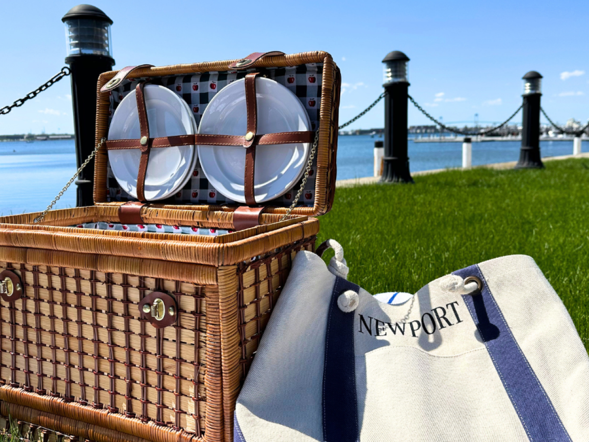 A picnic basket and a "Newport" tote bag are on grass by the water, with a fence and lampposts in the background.