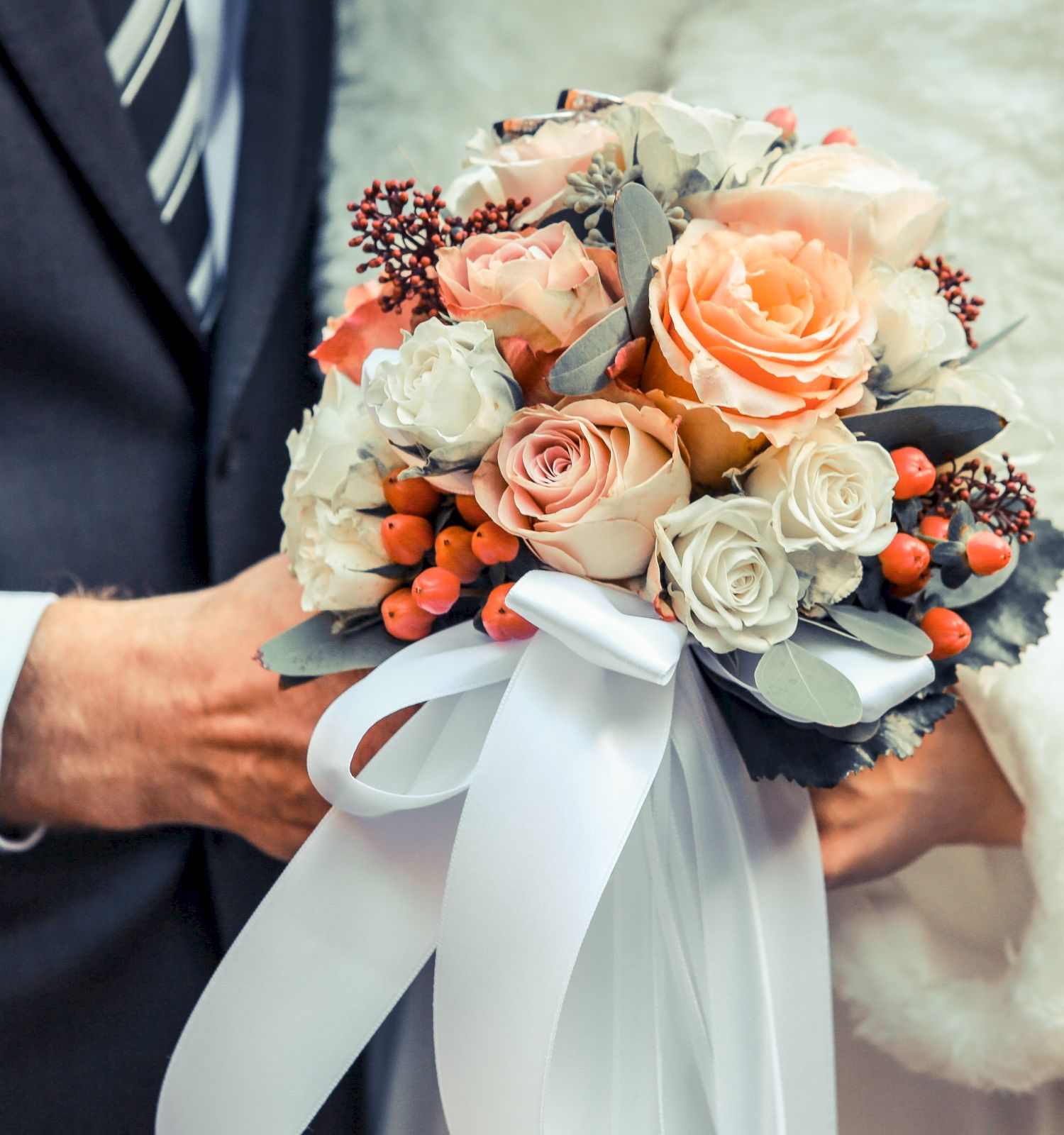 A couple holds a floral bouquet, featuring roses and berries, adorned with a white ribbon. One wears a suit, the other a white garment.