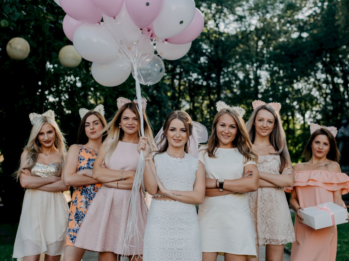 A group of women in dresses standing outdoors holding pink and white balloons, with trees in the background.