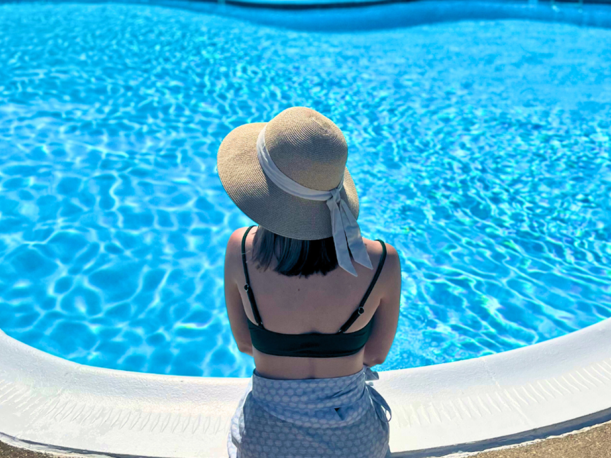 A person in a sun hat and swimsuit sits by the edge of a bright blue swimming pool, enjoying a sunny day.