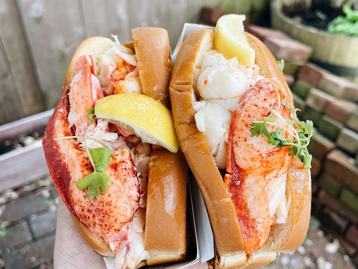 The image shows two lobster rolls garnished with lemon wedges and herbs, being held in a hand over a brick surface.