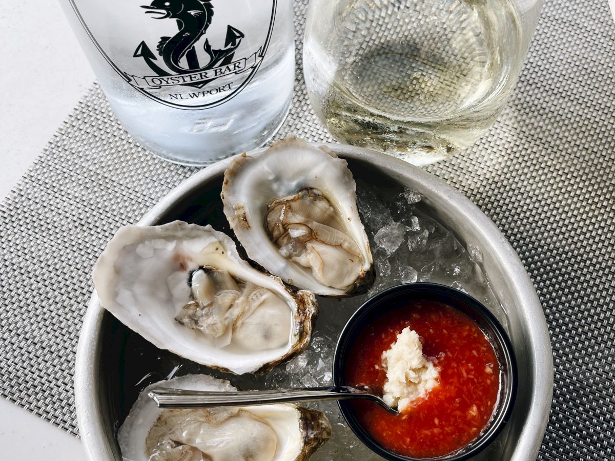 A dish of oysters on ice with cocktail sauce and lemon, a glass of white wine, and a glass of water with "MIDTOWN" branding visible.