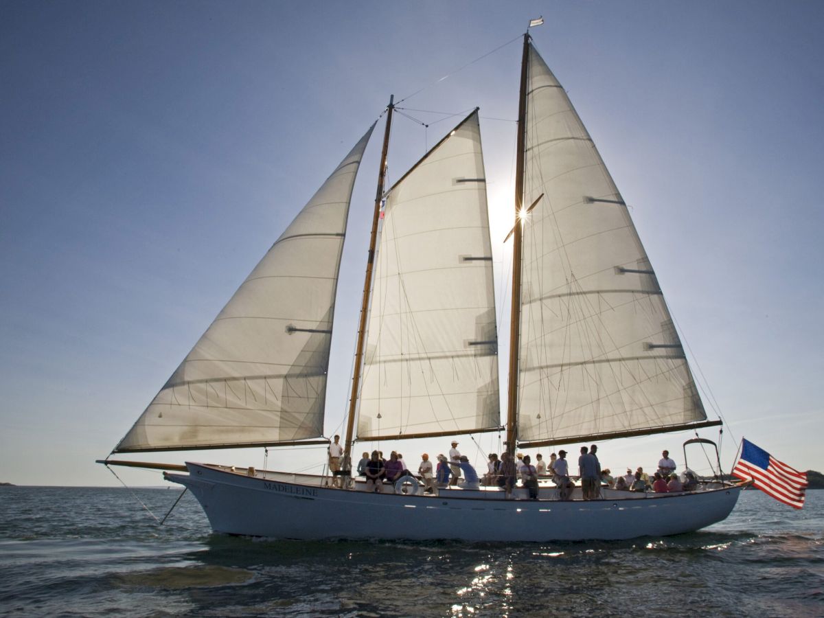 A sailboat with three sails navigates on the water with passengers, flying an American flag at the stern.