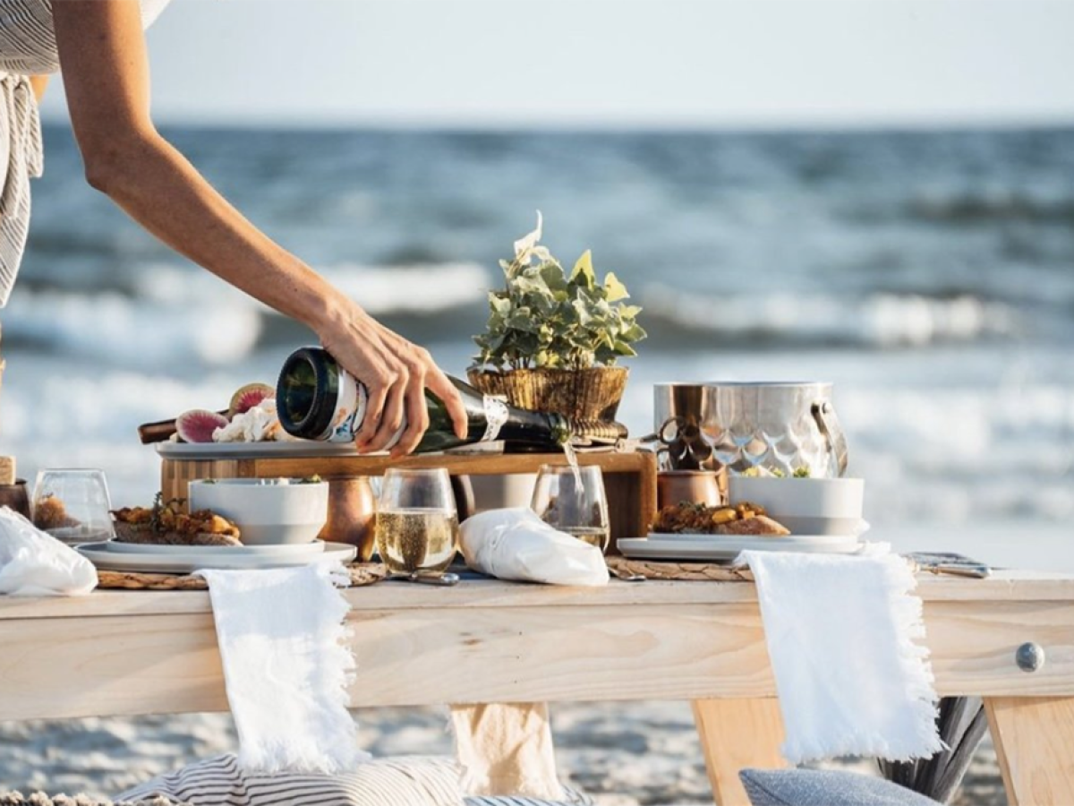 A beachside picnic setup features food, wine, and tableware on a wooden table, with someone pouring a drink by the sea.