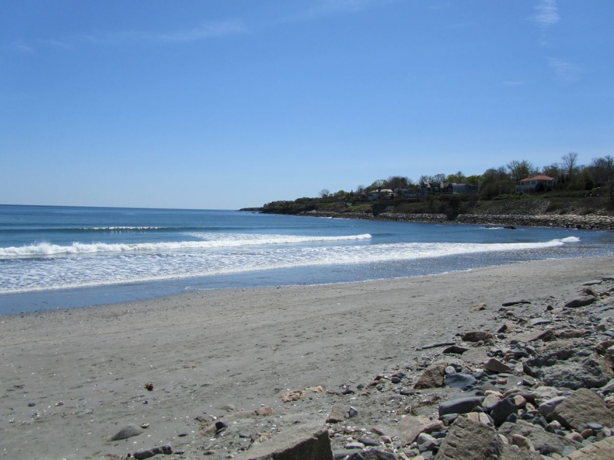A serene beach scene with gentle waves, a sandy shore, and rocky edges under a clear blue sky. Houses line the distant coast.