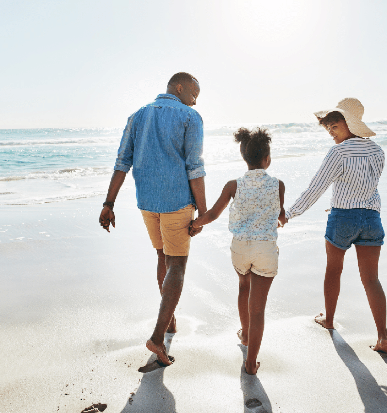 A family holding hands and walking along a sunny beach, with the ocean waves in the background.