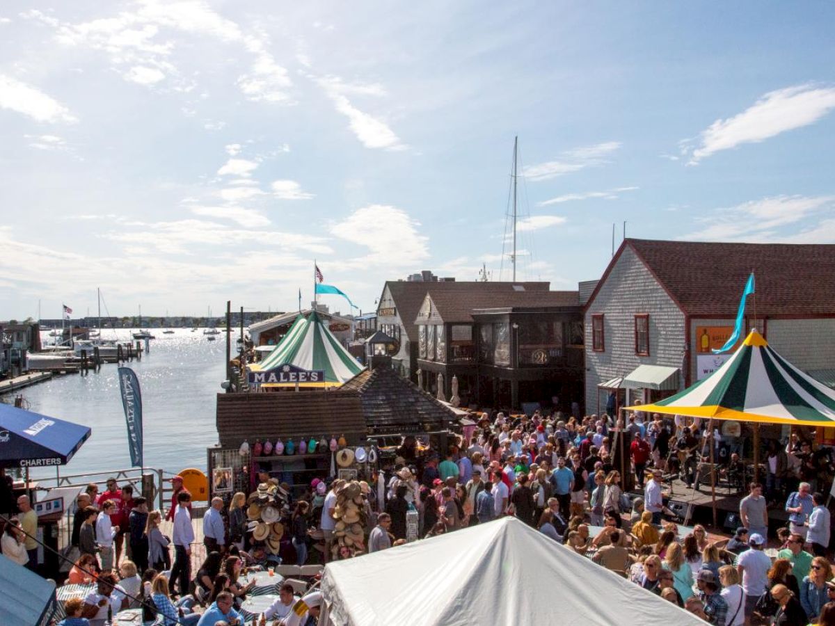 A bustling waterfront scene with tents, crowds, and boats moored, suggesting a lively outdoor market or festival atmosphere.