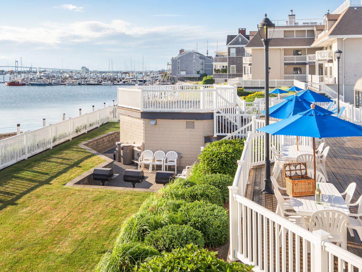 A waterfront patio with white railings and chairs, blue umbrellas, and lush greenery overlooks a marina and calm waters, with buildings nearby.