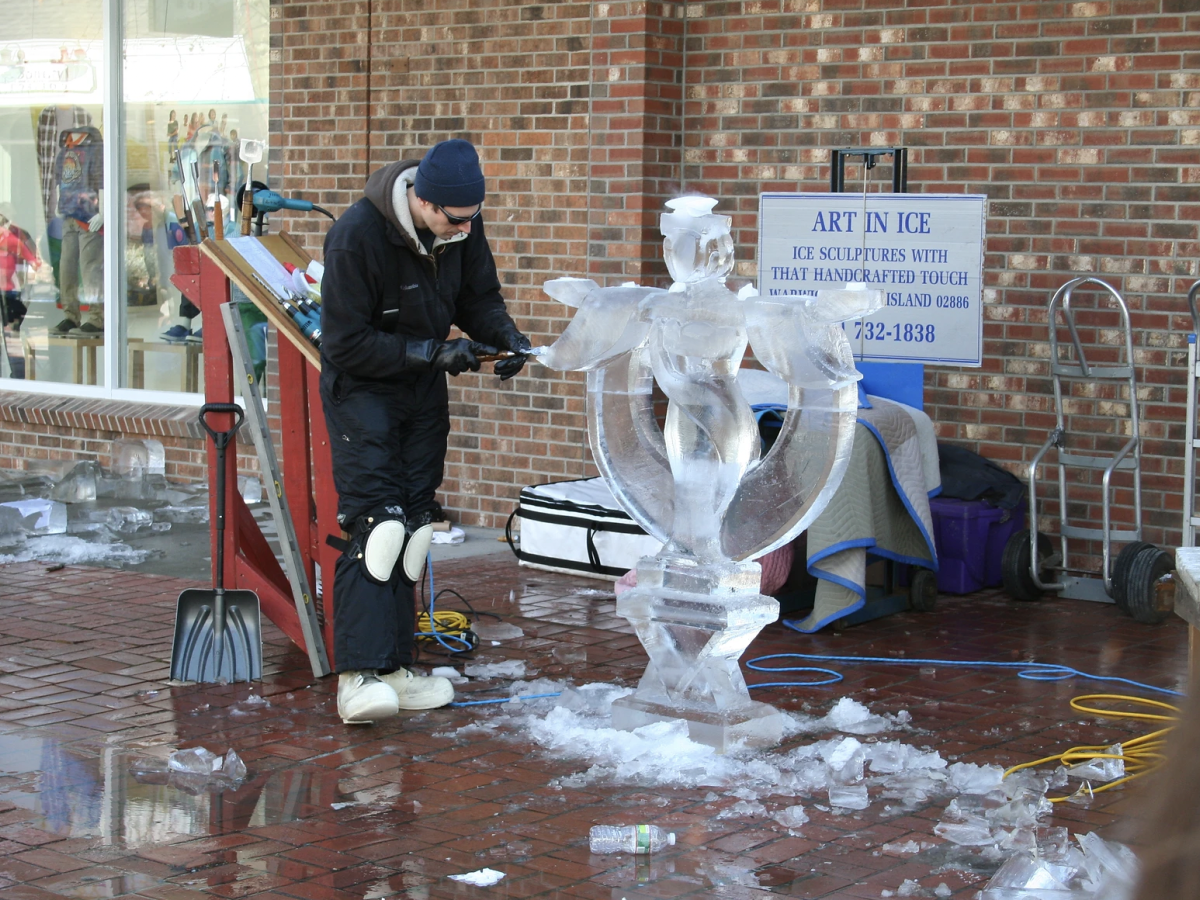 An artist is carving an ice sculpture outside, with tools scattered around. A sign behind them reads "Art in Ice" with a contact number.