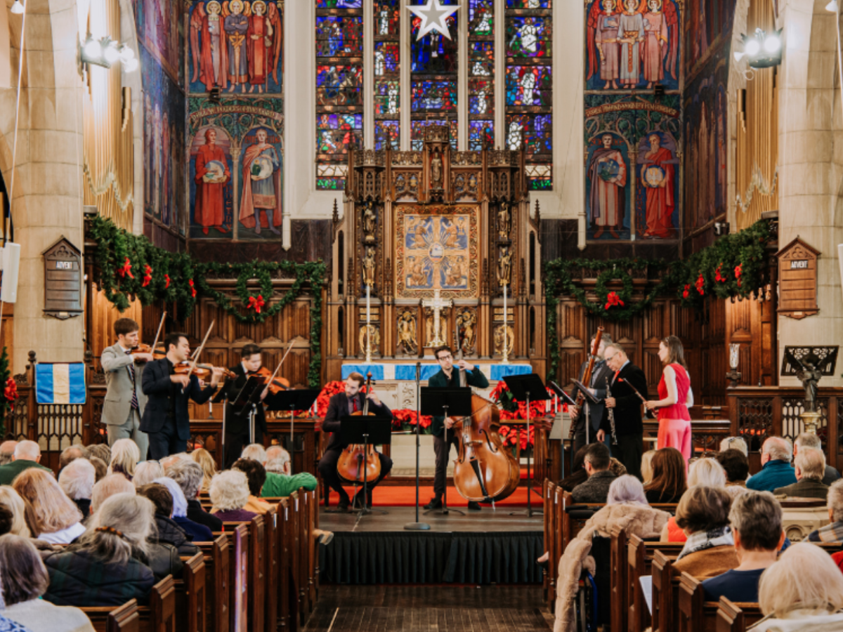 Musicians perform on a stage in a church with an audience seated in pews, surrounded by stained glass and decorative elements.