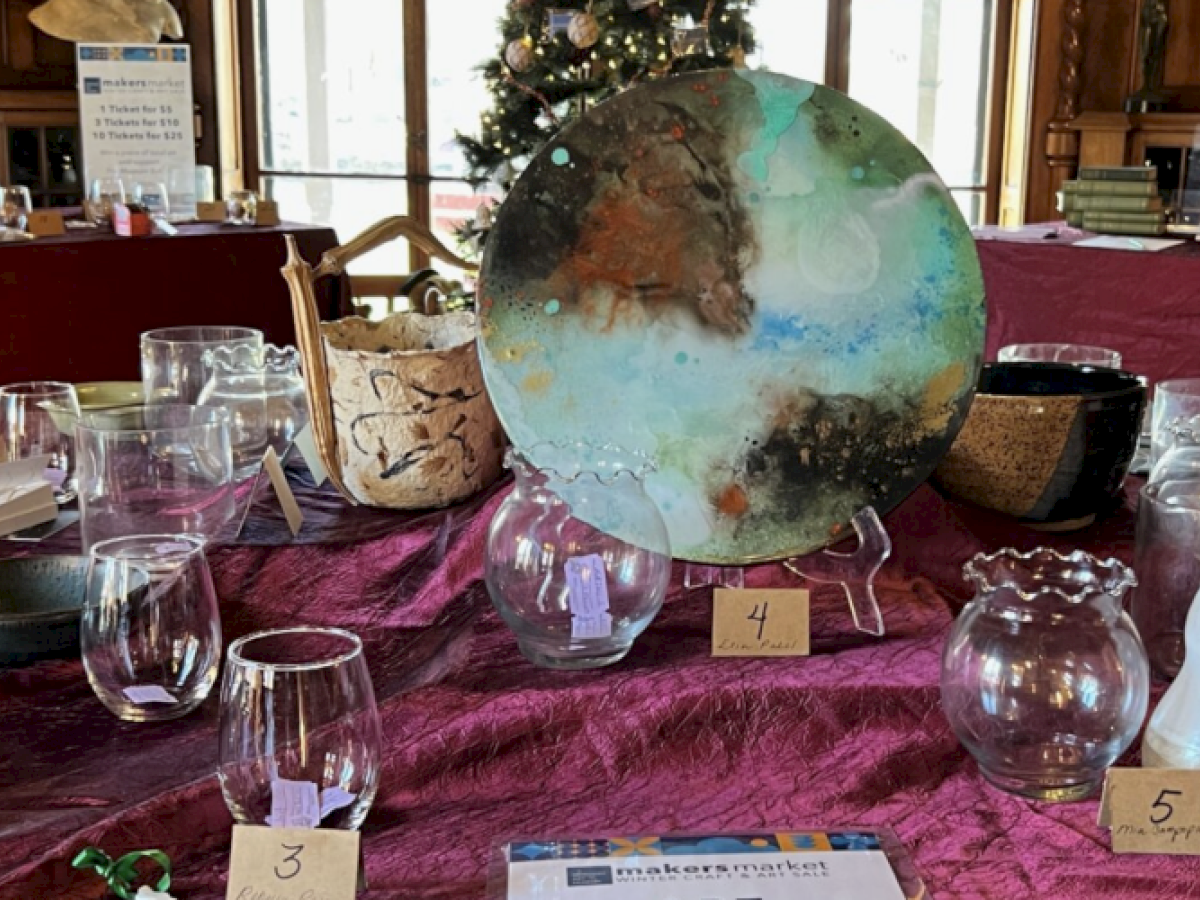 Glassware and decorative items are displayed on a table with number tags. A large colorful plate is at the center with a Christmas tree behind.
