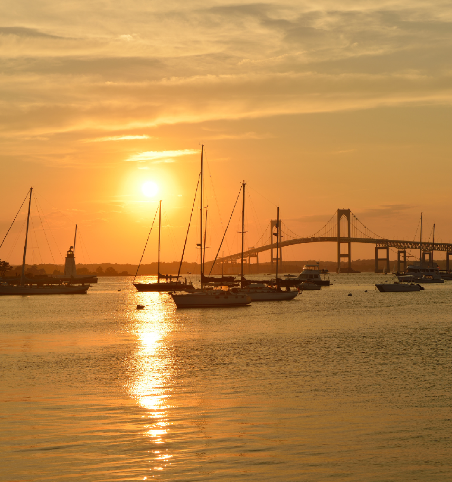 A tranquil sunset over water with sailboats and a bridge silhouetted against an orange sky, reflecting warmly on the water's surface.