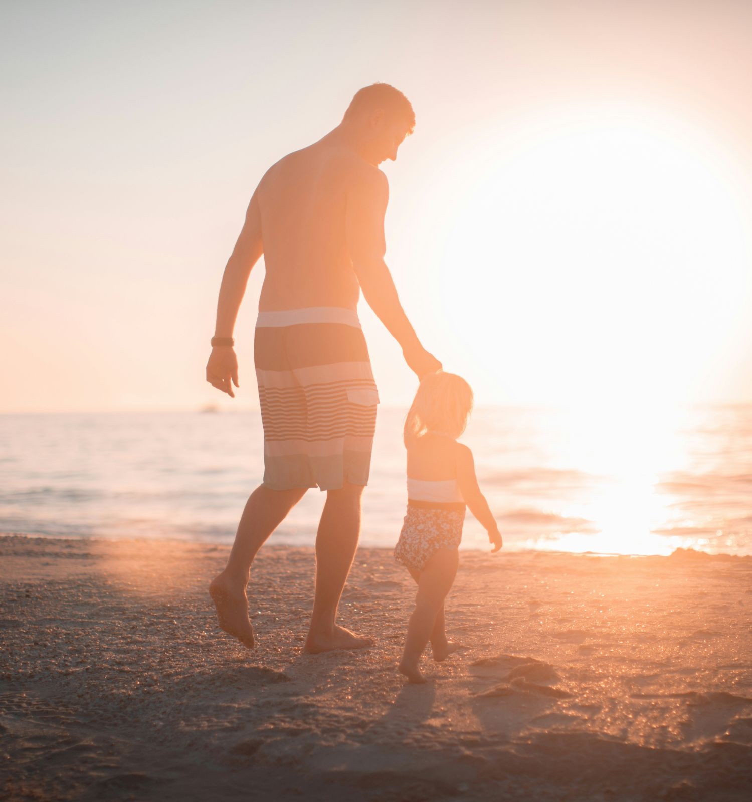A father and child walk hand-in-hand on a beach at sunset, silhouettes glowing as the sun dips toward the water.