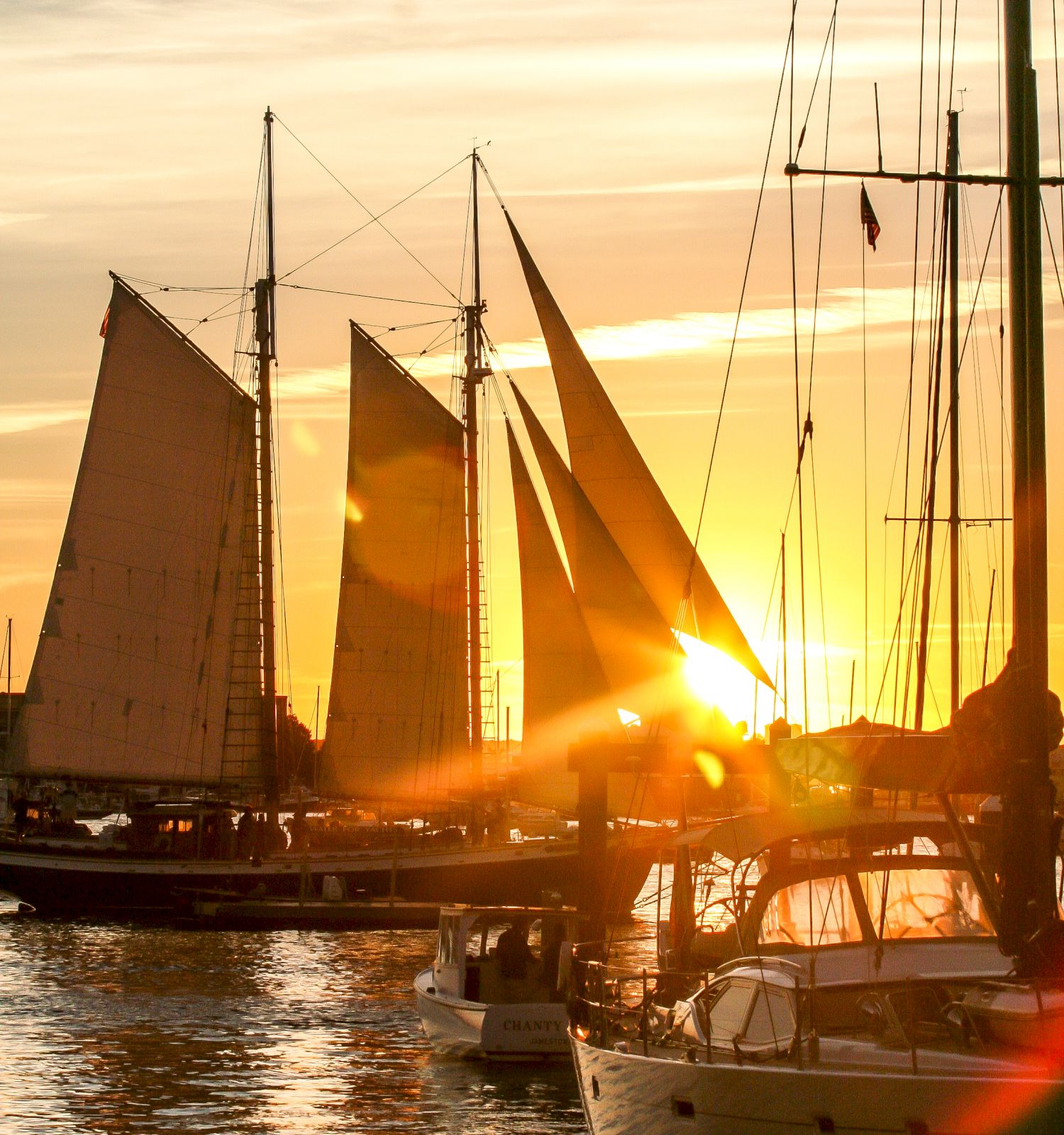 A sailing ship with tall rigging and foils is docked at sunset, silhouetted against the golden sky and reflected water.