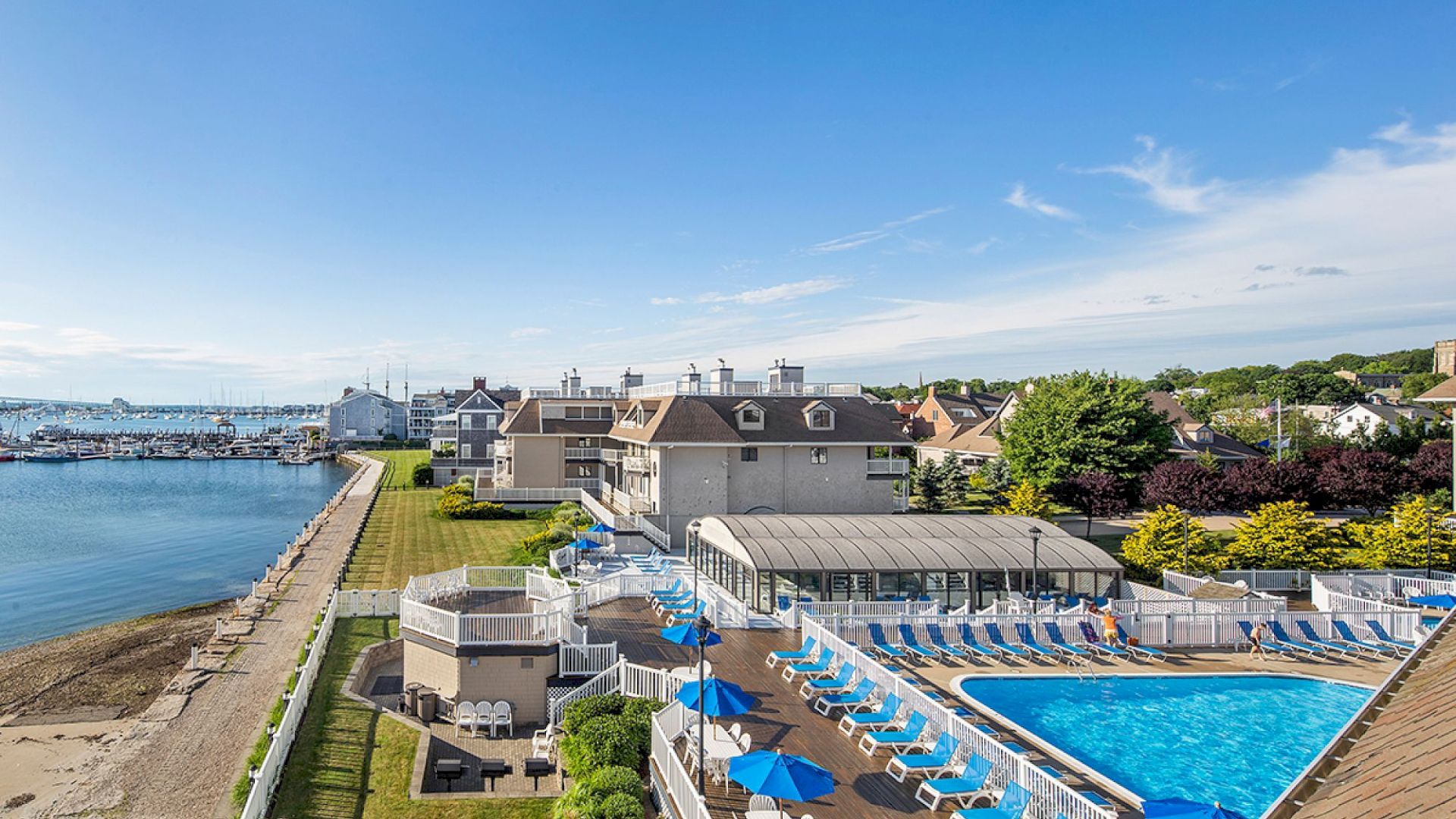 A seaside resort with a pool, sun loungers, and umbrellas next to a beach and marina under a clear blue sky.