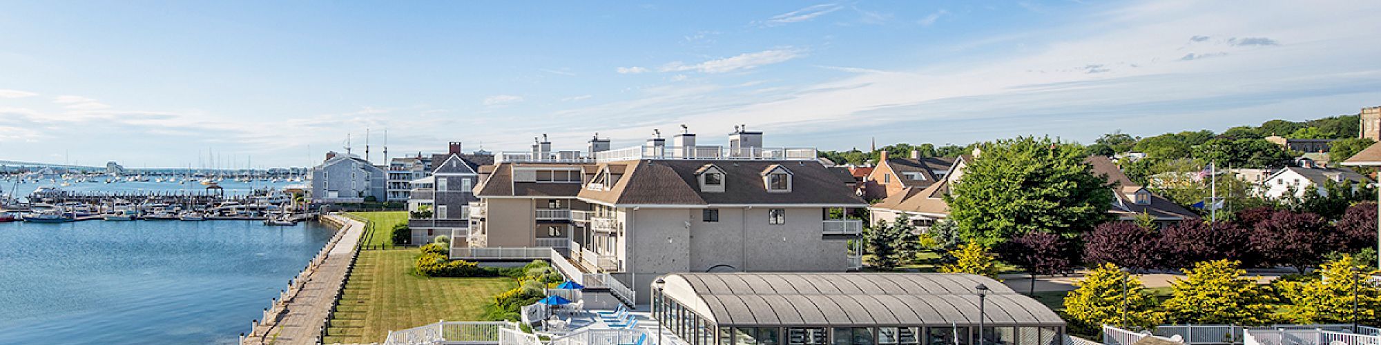 A seaside resort with a pool, sun loungers, and umbrellas next to a beach and marina under a clear blue sky.