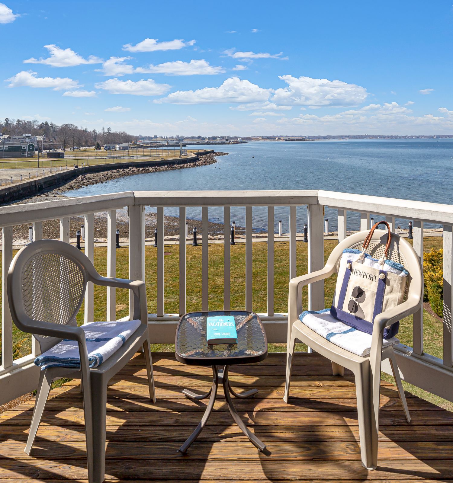 A waterfront deck with two chairs, a table holding a book and bag, surrounded by scenic views of water and sky.