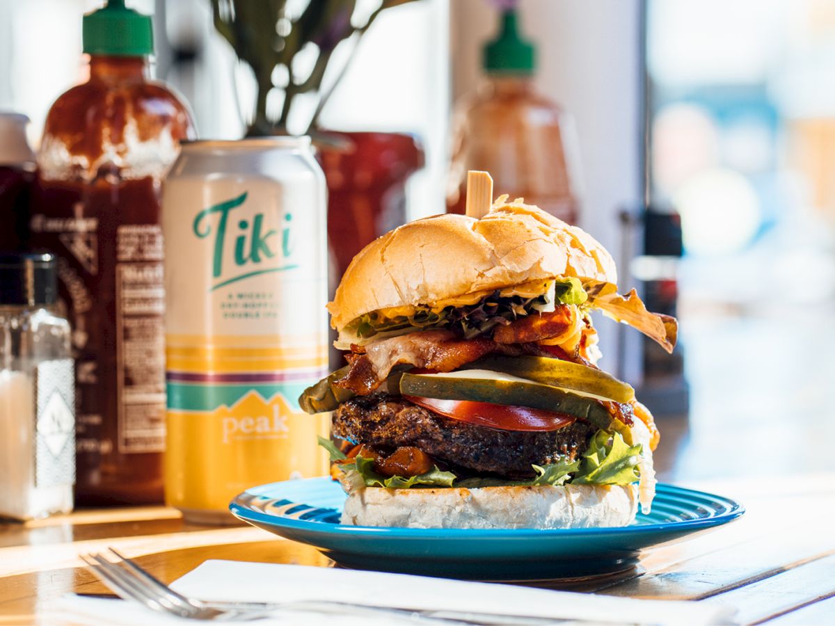 A loaded burger with toppings like bacon and pickles sits on a blue plate, with a Tiki drink can and condiments in the background.